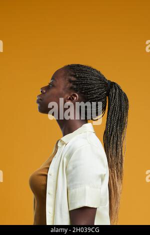 Ritratto minimal side view della giovane donna afroamericana in piedi su sfondo giallo in studio con focus sul lungo acconciatura delle trecce afroamericane Foto Stock