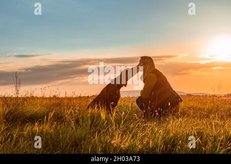 Donna matura e la sua bella vizsla ungherese. Amore del cane e background di amicizia. Donna che bacia il suo cane in una serata autunnale soleggiata. Foto Stock