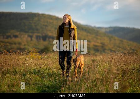 Donna matura che cammina la sua bella vizsla ungherese. Cane a piedi sfondo. Donna e cane da caccia godersi la natura a piedi in una soleggiata serata autunnale. Foto Stock