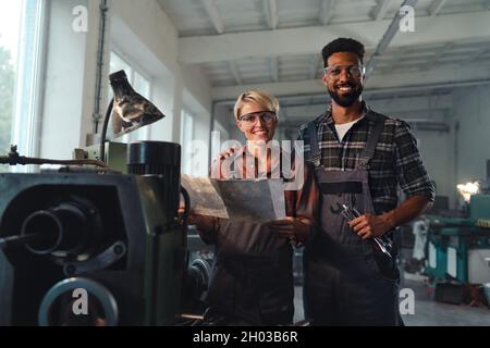 Ritratto di giovani colleghi industriali biraciali che lavorano in ambienti interni in un laboratorio di metallo, sorridendo e guardando la macchina fotografica. Foto Stock