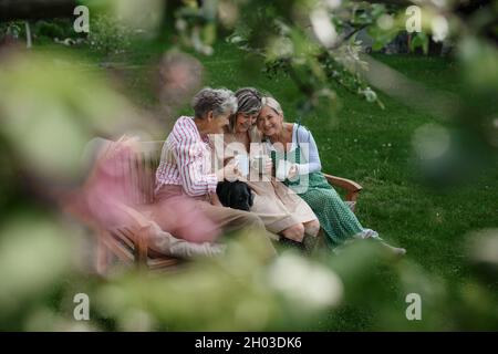 Felice donne anziane amici seduti su panca e bere tè all'aperto in giardino, ridendo. Foto Stock