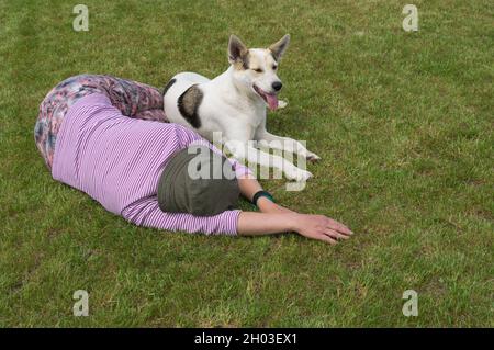 Donna caucasica matura e giovane cane bianco misto di razza adagiato uno accanto all'altro su un prato tagliato nella stagione estiva Foto Stock