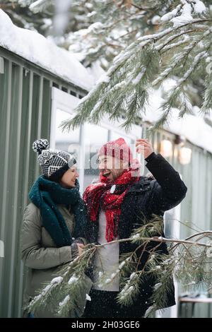 Coppia allegra che parla, ottenendo la neve dappertutto loro stessi dagli alberi vicini. Entrambi indossano cappelli, giacche e sciarpe. Di fronte ad una casa con corrugato Foto Stock