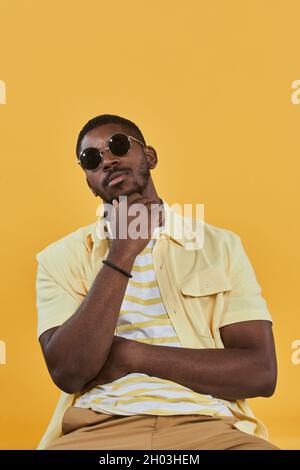 Ritratto verticale di un bell'uomo afro-americano che indossa occhiali da sole e guarda la macchina fotografica mentre si posa su sfondo giallo in studio Foto Stock
