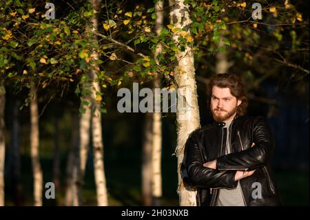 un giovane uomo barbuto in piedi accanto agli alberi nel parco autunnale e guardando attentamente in lontananza Foto Stock
