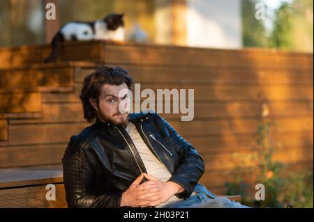 un giovane uomo in una giacca di pelle nera seduta sul terrazzo della casa e guardando con attenzione la macchina fotografica Foto Stock