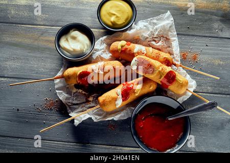 Vista dall'alto di deliziose salsicce in pasta di mais su spiedini posti su tavola di legno con salse assortite Foto Stock