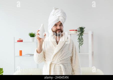 Ritratto di bellezza di bel ragazzo in asciugamano e accappatoio che tiene in mano il tubo di crema corpo lozione. Spa, cura del corpo e della pelle per l'uomo concetto Foto Stock