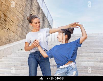 Madre di mezza età e sua figlia capretto in piedi su scale di pietra all'aperto, tenendo le mani e guardandosi l'un l'altro. Famiglia amorevole che indossa le t-shirt blu un Foto Stock