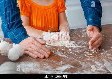 Piccola ragazza cucinando cibo in cucina con papà. Le mani di Daddy aiutano i bambini a impastare l'impasto. Figlia del bambino con impasto di farina per impastare. Padre Foto Stock