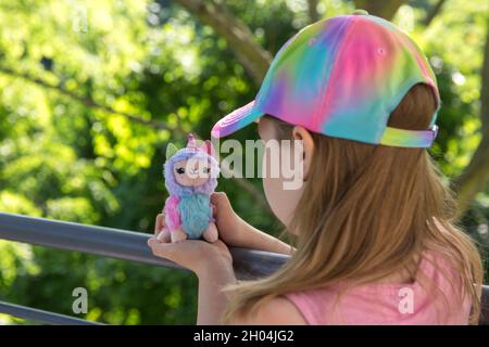 Unicorno di lama peluche colorata in mani bambina con green sullo sfondo. Famoso e popolare giocattolo per bambini. Foto Stock