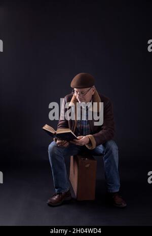 Uomo anziano seduto su valigia vecchia e libro di lettura, su sfondo nero Foto Stock