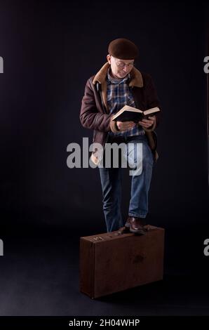 Uomo anziano in piedi accanto alla vecchia valigia e libro di lettura, su sfondo nero Foto Stock