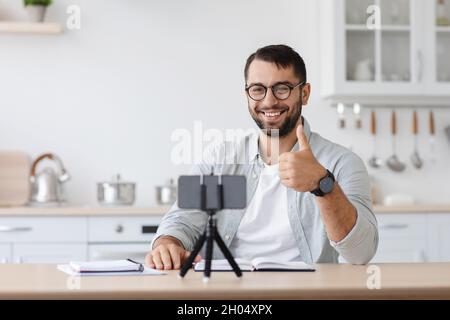 Felice adulto europeo maschio blogger con barba in occhiali in streaming video e mostrare pollici in su in cucina scandinava interni. Tutoraggio, studio, moderno r Foto Stock