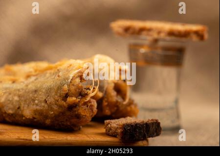 Un piccolo pesce fritto su un asse di legno, pezzi di pane e un bicchiere di vodka, primo piano, fuoco selettivo Foto Stock