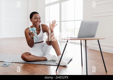 Eccitata atletica femmina nera in sportswear ondeggiante alla webcam facendo video conferenza con personal trainer o follower, seduto sul pavimento su yoga Foto Stock