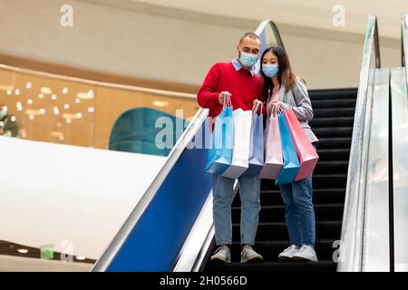 Tutta la lunghezza di giovane coppia multirazziale in maschere di faccia scendendo sulla scala mobile al centro commerciale, guardando in borse shopping, spazio copia. Coniuge millenario Foto Stock