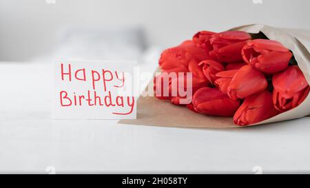 Buon compleanno. Bouquet di tulipani rossi e biglietto d'auguri con iscrizione scritta a mano buon compleanno su sfondo bianco. Fiori anniversario della donna Foto Stock