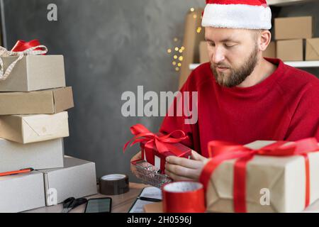 Ritratto di un uomo d'affari in un cappello di Santa che lavora in un ufficio domestico o magazzino con le scatole dei regali per Natale. Un uomo mette la scatola in un cestino di shopping Foto Stock