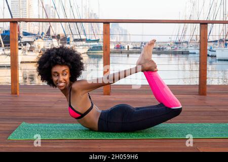 Giovane atletica in abbigliamento sportivo che si estende su un tappeto su un ponte di legno in un porticciolo Foto Stock