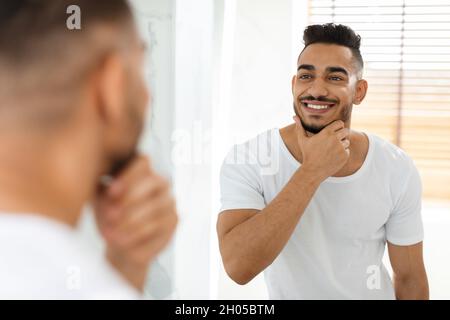 Cura del viso maschile. Bell'uomo Arabo che guarda nello specchio in bagno e toccando barba, attraente Guy Masculine Medio Oriente sorridente a Reflection, E. Foto Stock