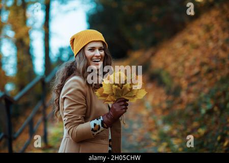 Ciao autunno. Felice elegante donna di 40 anni in cappotto marrone e cappello giallo con foglie giallo autunno a piedi tour all'aperto nel parco della città in autunno Foto Stock
