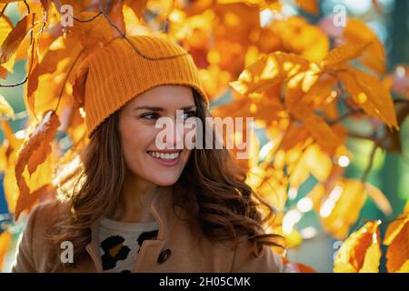 Ciao autunno. Felice donna moderna di 40 anni in cappotto beige e cappello arancione tra il fogliame autunnale fuori sul parco della città in autunno. Foto Stock