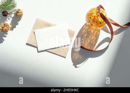 Composizione di Natale. Biglietto di auguri vuoto, busta mockup.decoration in luce solare.sfera di vetro di Natale, nastro marrone, rami di abete e cono di pino Foto Stock