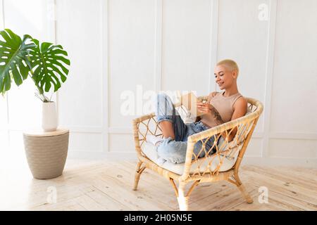 Concetto di attività del fine settimana. Donna rilassante in comoda poltrona in vimini con libro a casa, spazio copia. Signora felice prendendo pausa dal lavoro Foto Stock