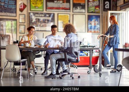 gruppo multietnico di colleghi che lavorano insieme, sorridenti, si siedono alla scrivania con il loro collega che si trova accanto a uno scooter elettrico, osservando Foto Stock