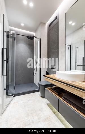 Moderno e minimalista bagno luminoso con mobili in legno, piastrelle in pietra grigia e beige e cabina doccia in vetro. Design degli interni Foto Stock