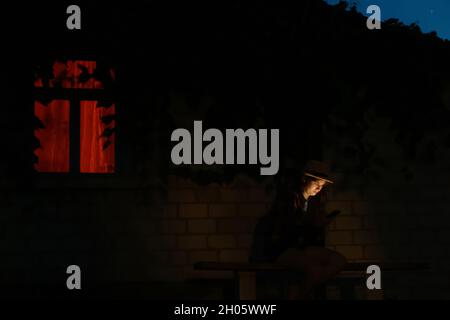 Defocalizzare la giovane donna utilizzando il sedile per smartphone sulla panca di notte vicino a casa. Ritratto all'aperto di millennial usando il suo cellulare di notte. Arancione ligh Foto Stock