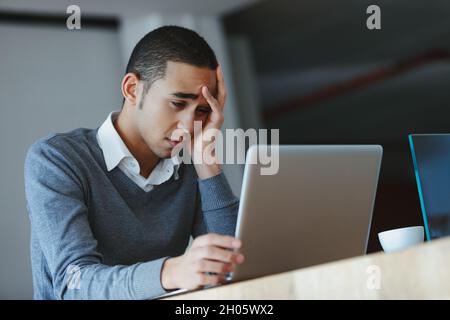 Annoiato, preoccupato o esausto uomo d'affari che fissava il suo computer portatile con testa a portata di mano e un'espressione seria turbata come si siede a una scrivania Foto Stock