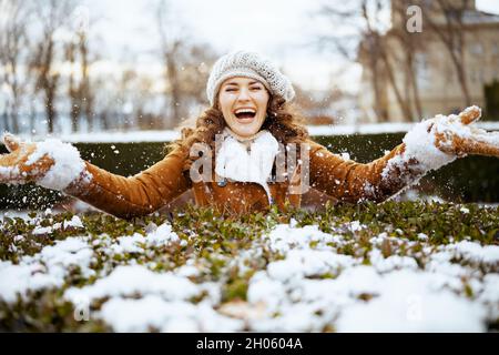 sorridente donna elegante con guanti in un cappello in maglia e cappotto di pelle di pecora gettando neve all'aperto nel parco della città in inverno. Foto Stock