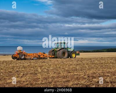 Un trattore verde John Deere che aratura un campo di stoppie in una giornata parzialmente soleggiata in autunno. Preso in Northumberland, Inghilterra, Regno Unito Foto Stock