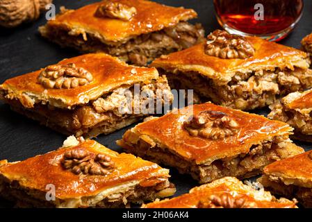 Miele baklava con noci su ardesia nera. Porzioni di dolci orientali. Pezzi zuccherini turchi. Tè versato in vetro di armudu sullo sfondo. Foto Stock
