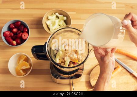 Mani di femmina che versano il latte nel frullatore elettrico con ingredienti tritati durante la preparazione del frullato Foto Stock