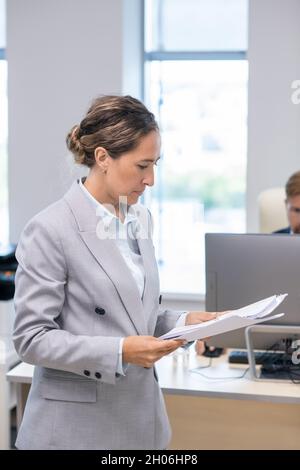 Giovane donna d'affari seria che guarda attraverso i documenti finanziari mentre lavora in ufficio Foto Stock