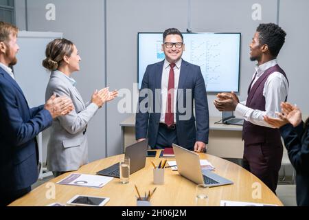 I dipendenti interculturali sono in piedi da un grande tavolo e si aggrappano le mani mentre si congratulano con il loro capo in abito Foto Stock