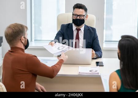 Uomo d'affari maturo in maschera protettiva che passa carta finanziaria al giovane durante la consultazione Foto Stock