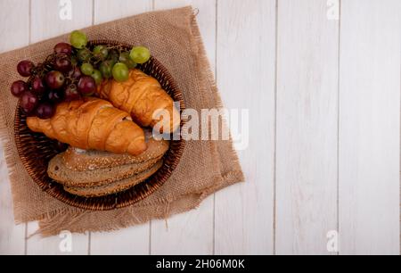 vista dall'alto di pane come croissant e fette di pane di panino bruno seminato con uva nel cestino su sackcloth su sfondo di legno con spazio copia Foto Stock