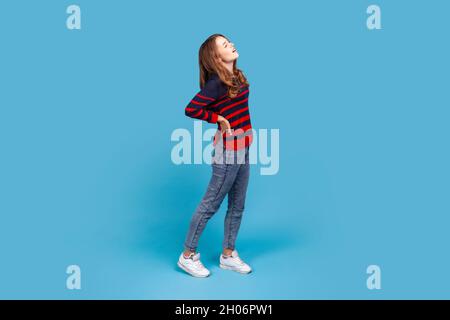 Tutta la lunghezza di donna che indossa un pullover casual a righe, con forte mal di schiena, toccando e premendo le mani alla schiena, chiudendo gli occhi per alleviare il dolore. Studio interno girato isolato su sfondo blu. Foto Stock