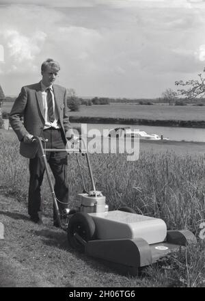 Anni '60, storico, fuori su terra erbosa accanto ad un fiume, un giovane uomo in una giacca sportiva a scacchi e cravatta, utilizzando un grande tosaerba a benzina gommato per tagliare l'erba lunga, Oxford, Inghilterra, Regno Unito Foto Stock