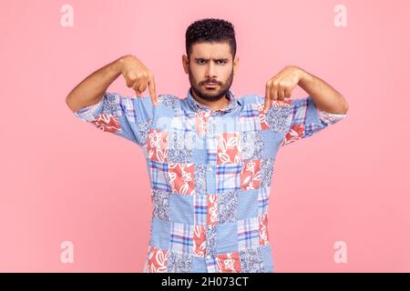 Qui e ora. Uomo stretto bearded bello che punta il dito verso il basso, guardando la macchina fotografica con l'espressione facciale grave bossy, indossando la camicia casual blu. Studio interno girato isolato su sfondo rosa. Foto Stock