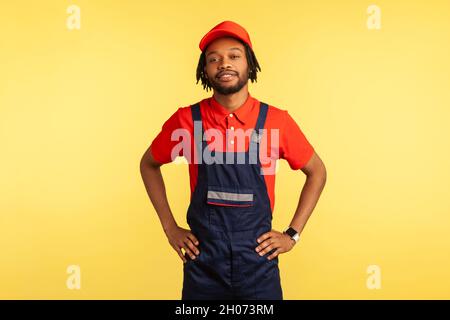 Ritratto di uomo di fiducia in piedi e tenere le mani in vita con sorriso, in attesa di ordini di lavoro, uomo di lavoro indossando tute, interni studio girato isolato su sfondo giallo. Foto Stock