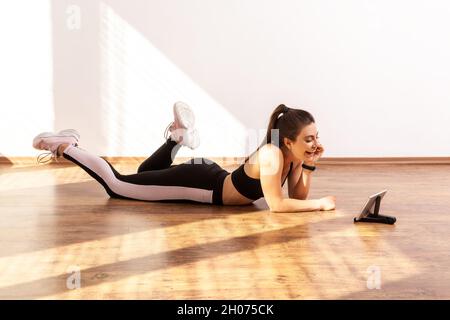 Donna snella che si trova sul pavimento e guarda il video sul fitness che si esercita a casa, sembra felice, indossando il top sportivo nero e tights. Ripresa completa in studio illuminata dalla luce del sole dalla finestra. Foto Stock