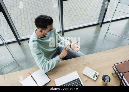 Vista dall'alto di un uomo d'affari arabo in maschera medica spray igienizzatore a mano vicino a caffè e computer portatile in ufficio Foto Stock