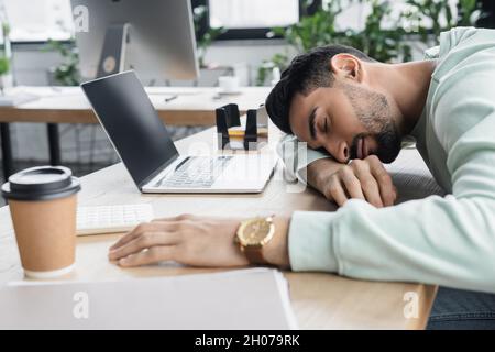 Giovane uomo d'affari arabo che dorme vicino al caffè per andare e al computer portatile in ufficio Foto Stock