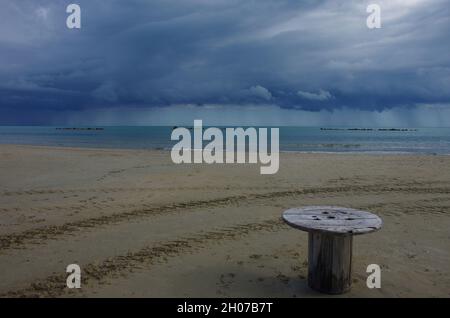 In primo piano sulla spiaggia deserta un tavolo fatto da una grande bobina di legno, sullo sfondo, sul mare si può vedere una tempesta che arriva. Foto Stock