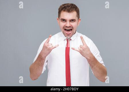 Ritratto di giovane uomo bello in camicia bianca e cravatta in piedi con canti di roccia e urla. Interno isolato su sfondo grigio. Foto Stock